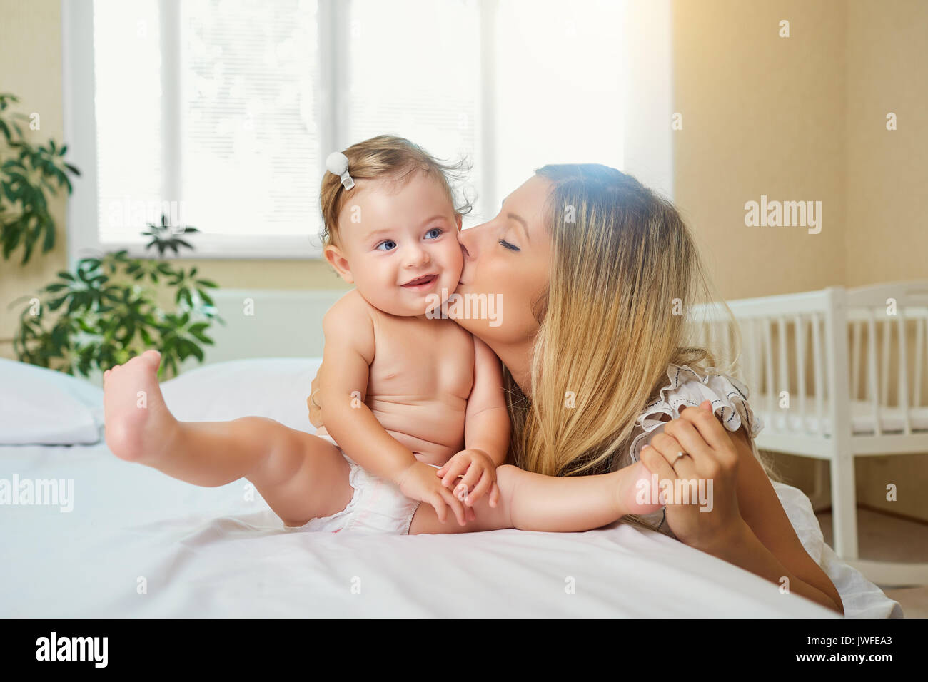 Mother and baby in a diaper play hugging on a bed indoors.  Stock Photo
