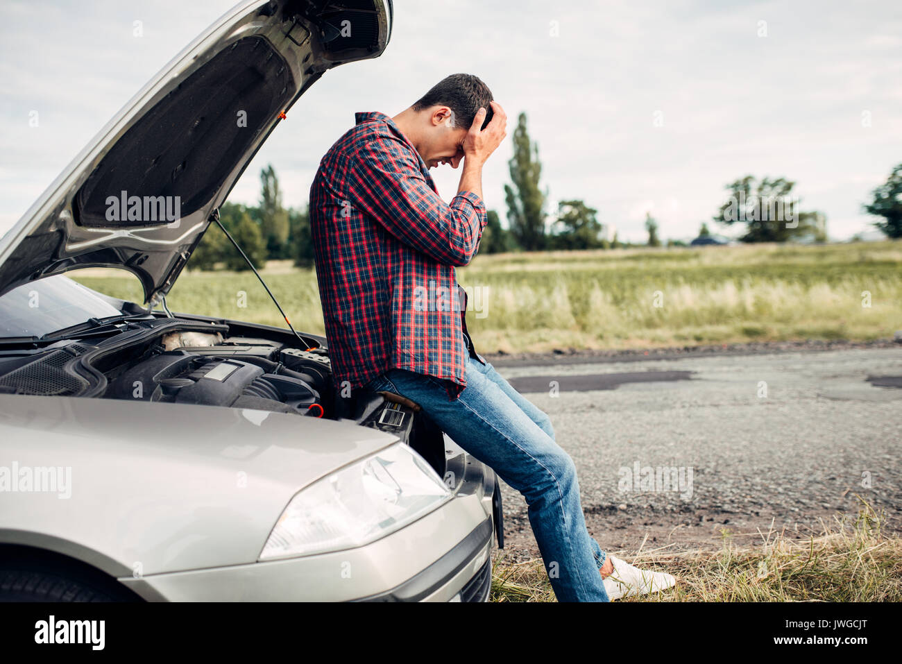 Depressed man sitting on a hood of broken car. Vehicle with open hood