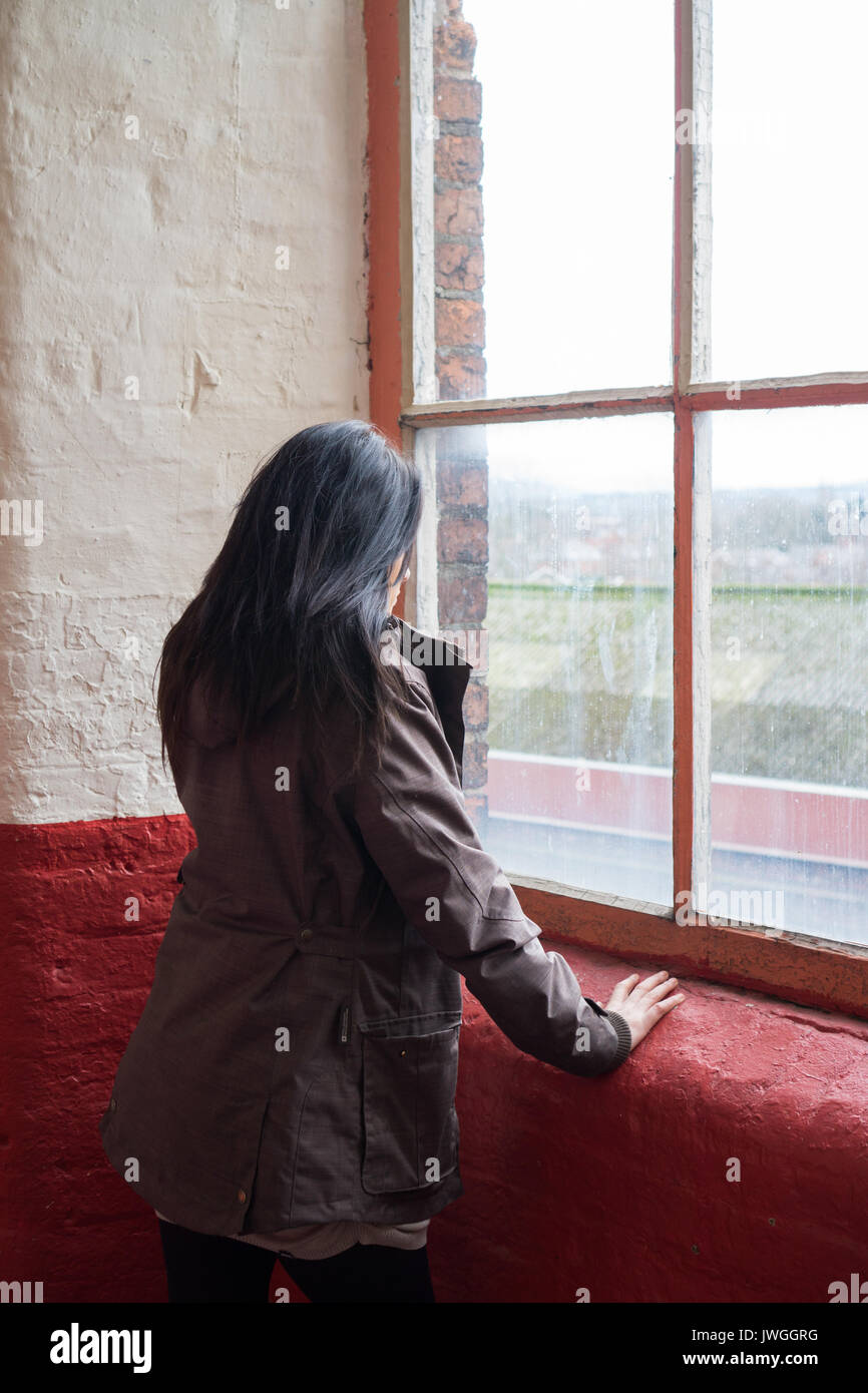 Rear view ,of a young woman with dark hair wearing a coat standing by the window Stock Photo