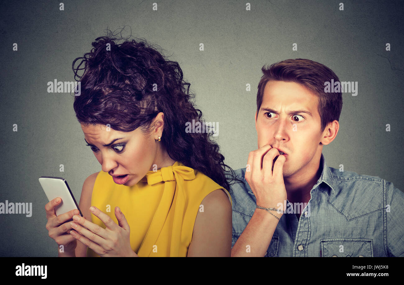 A cheating boyfriend. Anxious man nervously biting fingernails while shocked girlfriend reading text messages on his mobile phone Stock Photo