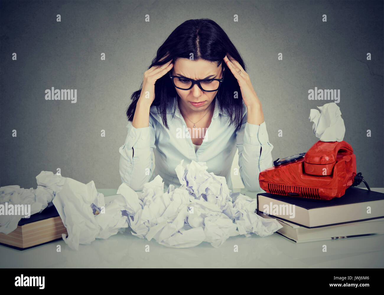 Too much work stressed woman sitting at her disorganized desk with books and many paper balls isolated on gray office wall background. Stock Photo
