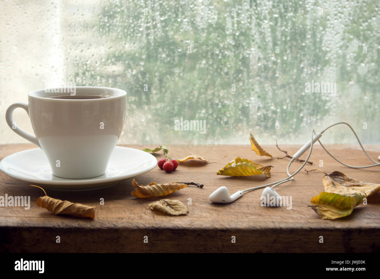 Cup of coffee or tea, smartphone and earbuds with autumn leaves near a window. Autumn playlist concept. Autumn music for rainy days. Stock Photo
