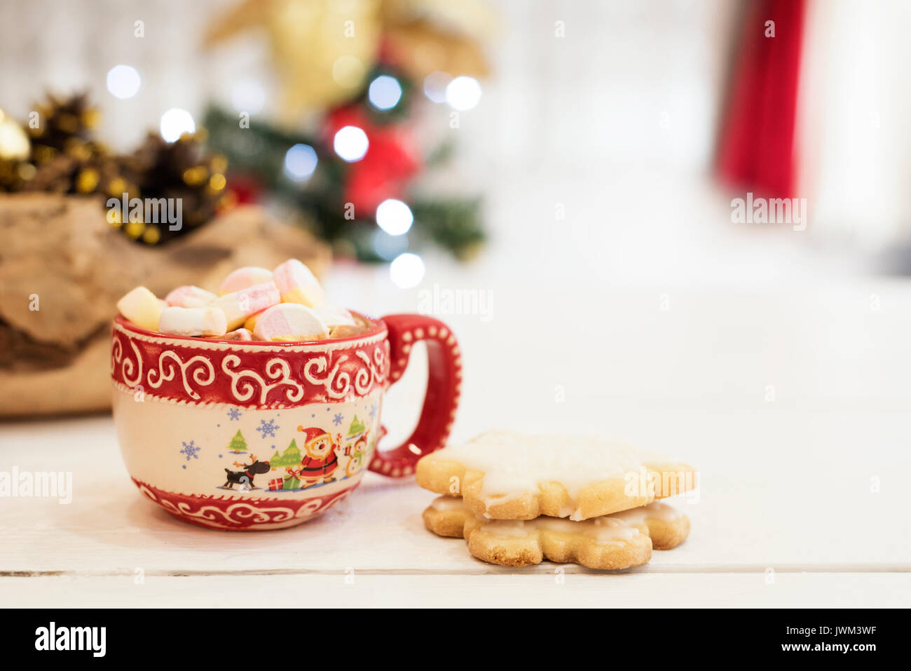 Hot chocolate with marshmallow candies. Christmas cookies shaped in snowflakes,  golden cones and christmass tree lights. White wooden background Stock Photo