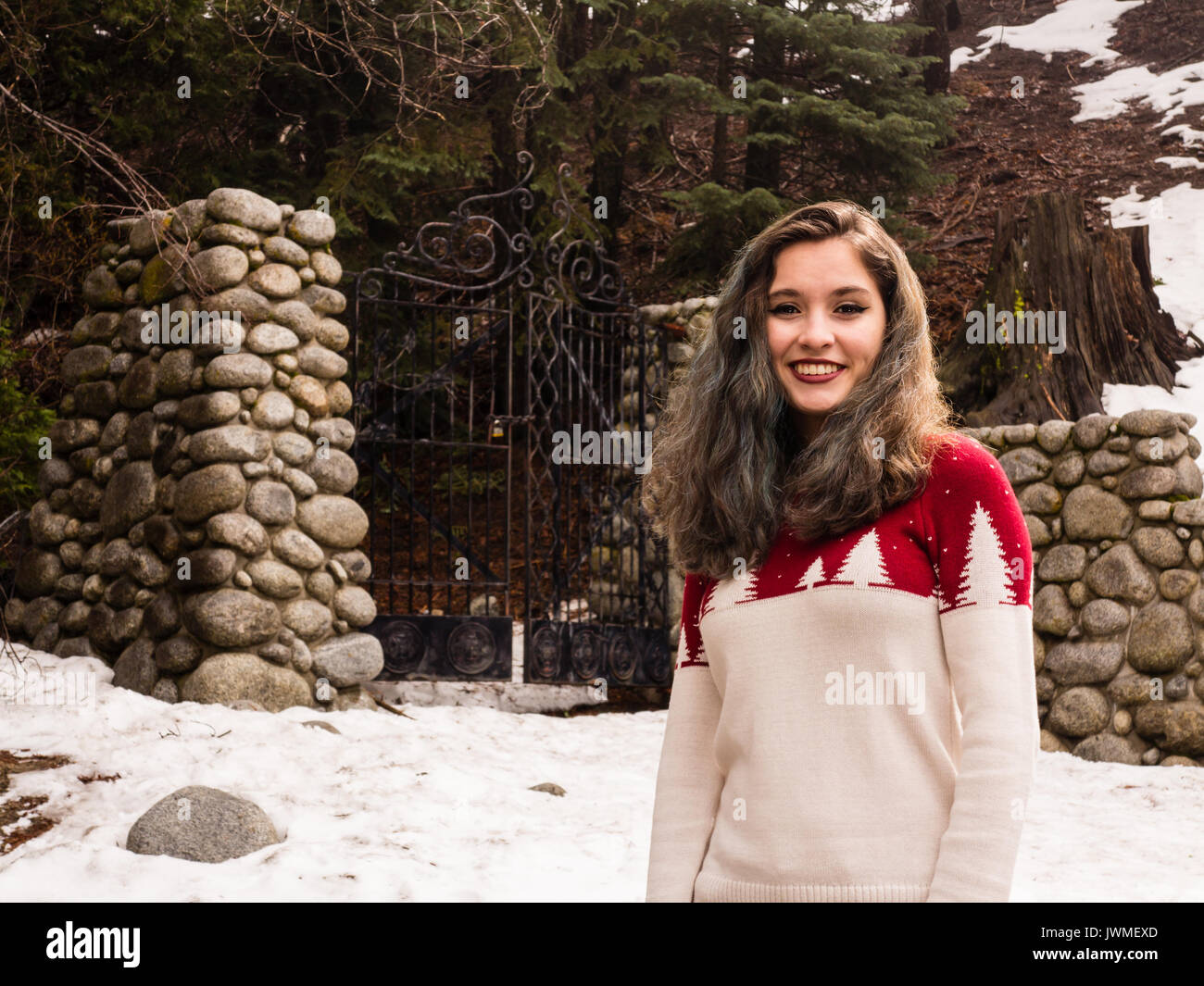 A smiling teen girl in red and white sweater by an iron gate with stone gateposts in winter Stock Photo