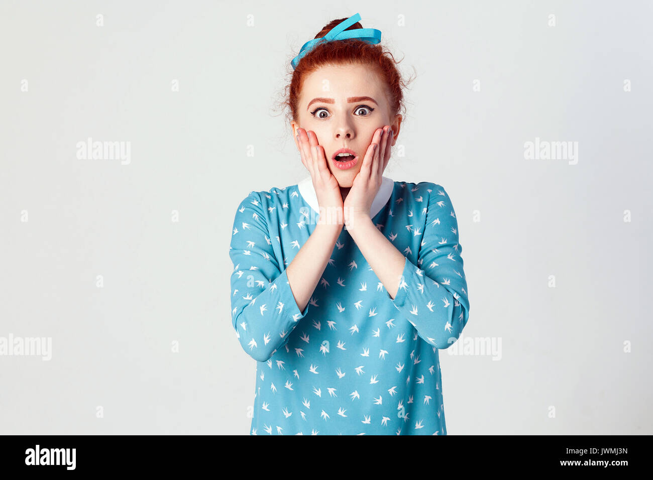 Human face expressions and emotions. Redhead young girl screaming with shock, holding hands on her cheeks. Isolated studio shot on gray background. Stock Photo