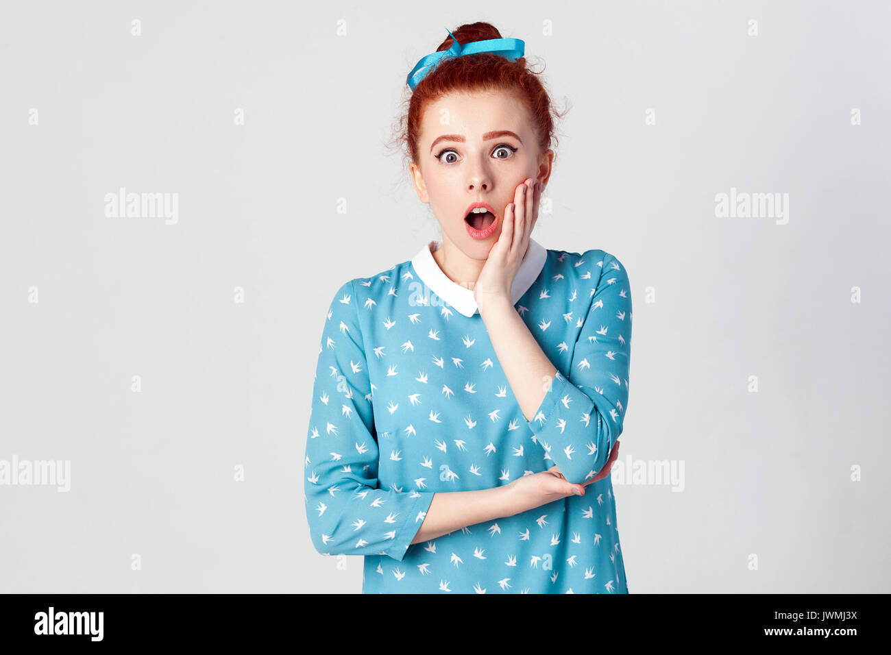 Ginger young girl screaming with shock, holding hands on her cheeks. Isolated studio shot on gray background. Stock Photo