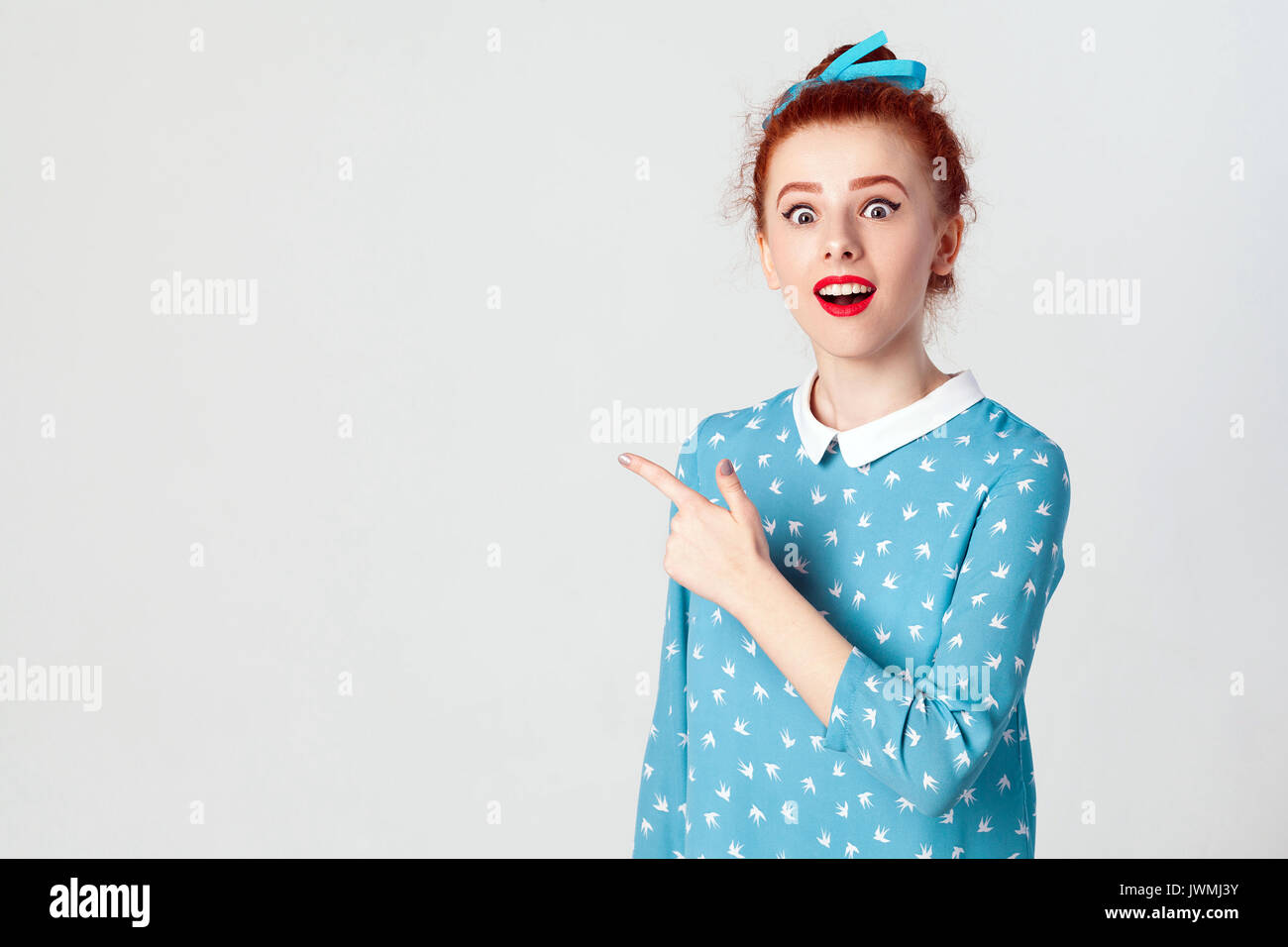 Excited young redhead caucasian girl with hair knot pointing her index finger sideways, raising eyebrows and keeping mouth wide opened, showing someth Stock Photo