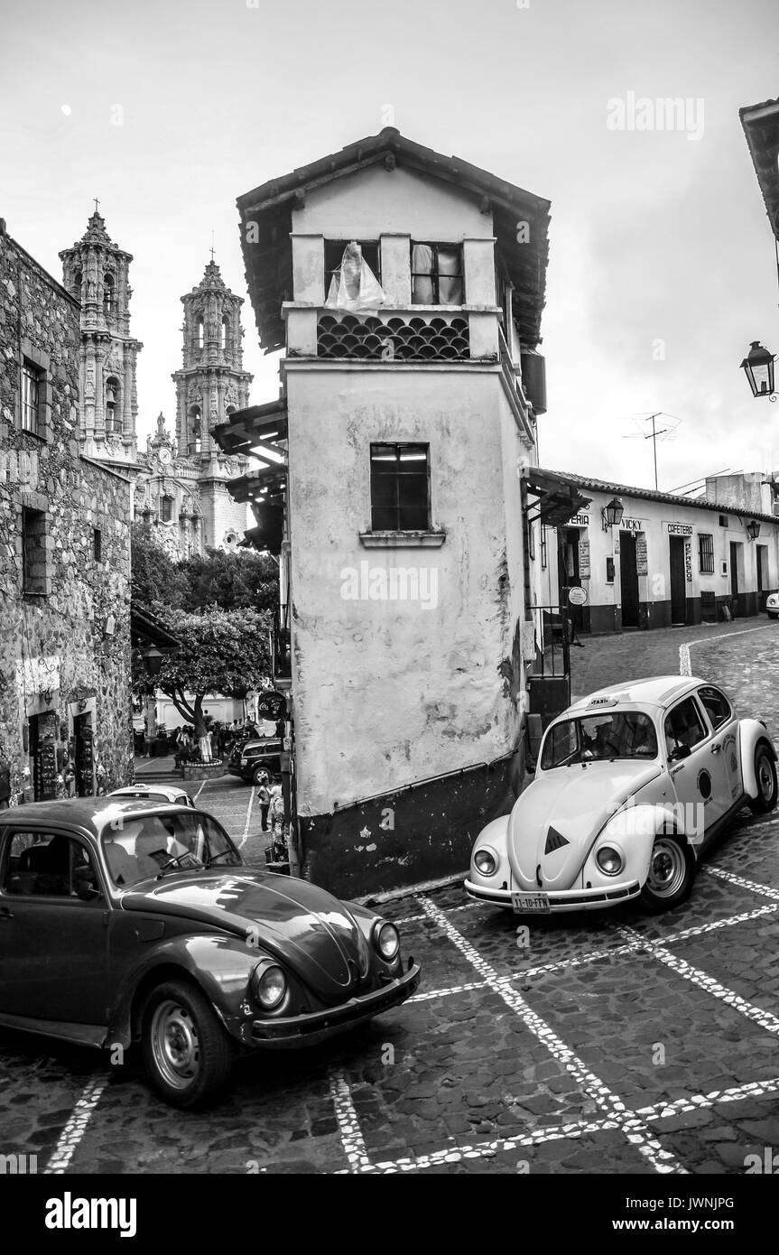 Two Volkswagen beetles negotiating a tight sloped bend around historic buildings in Taxco, southern Mexico, black and white image Stock Photo