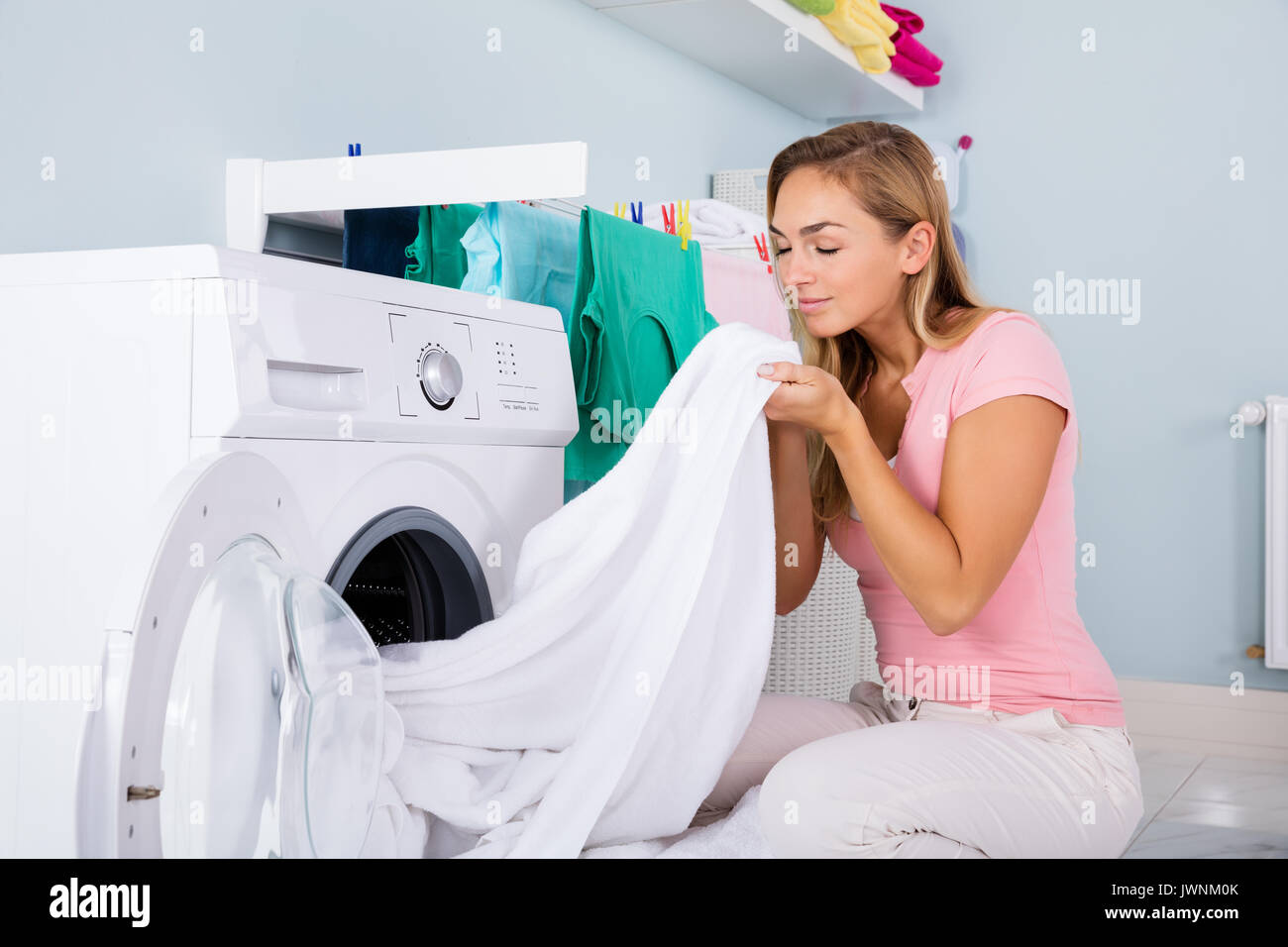Young Woman Smelling Clean Clothes Near The Electronic Washer At Laundry Room Stock Photo