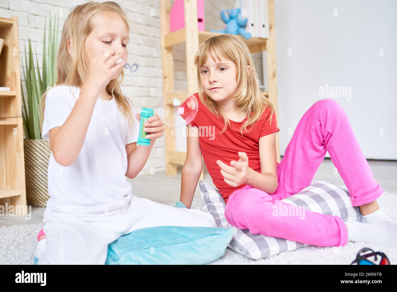 Pretty blond-haired sisters wearing home clothing gathered together in cozy living room and blowing bubbles Stock Photo
