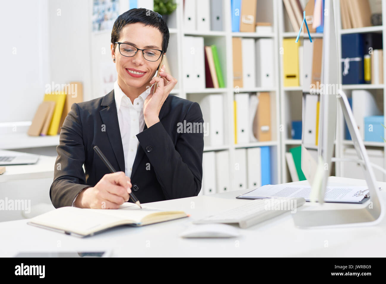 Attractive middle-aged designer writing down order details while talking to her client on mobile phone, interior of modern office on background Stock Photo