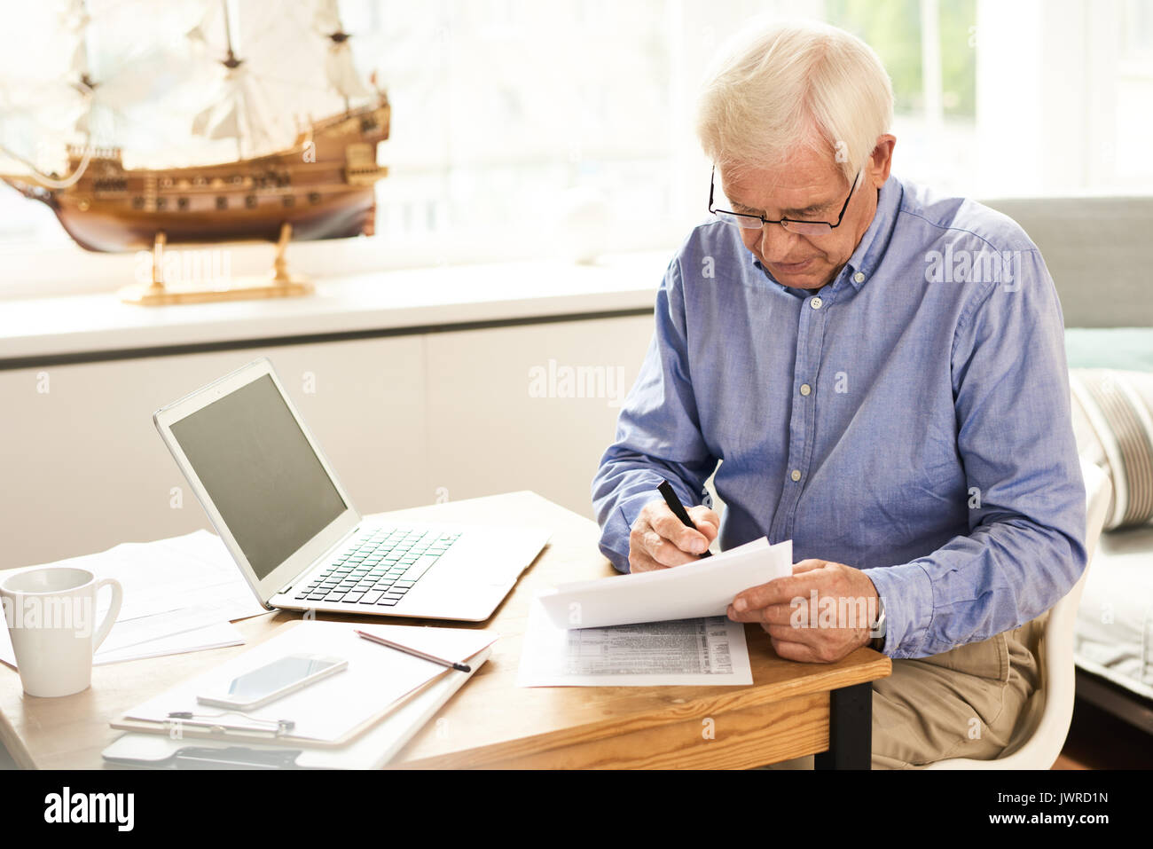 Portrait of modern senior man filling in application papers at home sitting at table with laptop in cozy living room Stock Photo