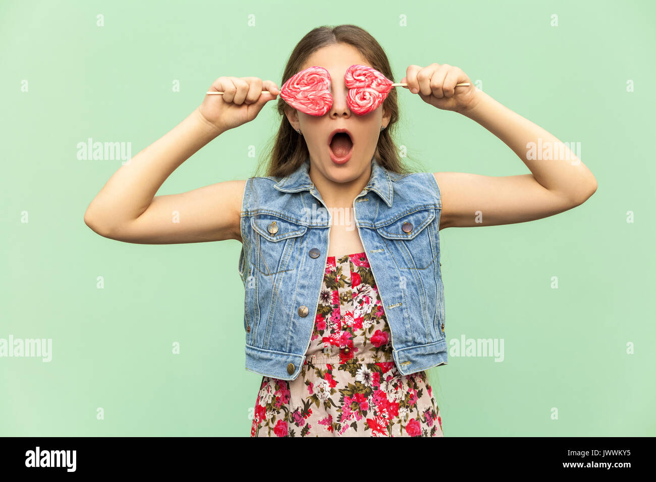 The teenager girl with blondie long hair, in shocked, holds two lolipop on her eyes, having funny look. Isolated studio shot on light green background Stock Photo