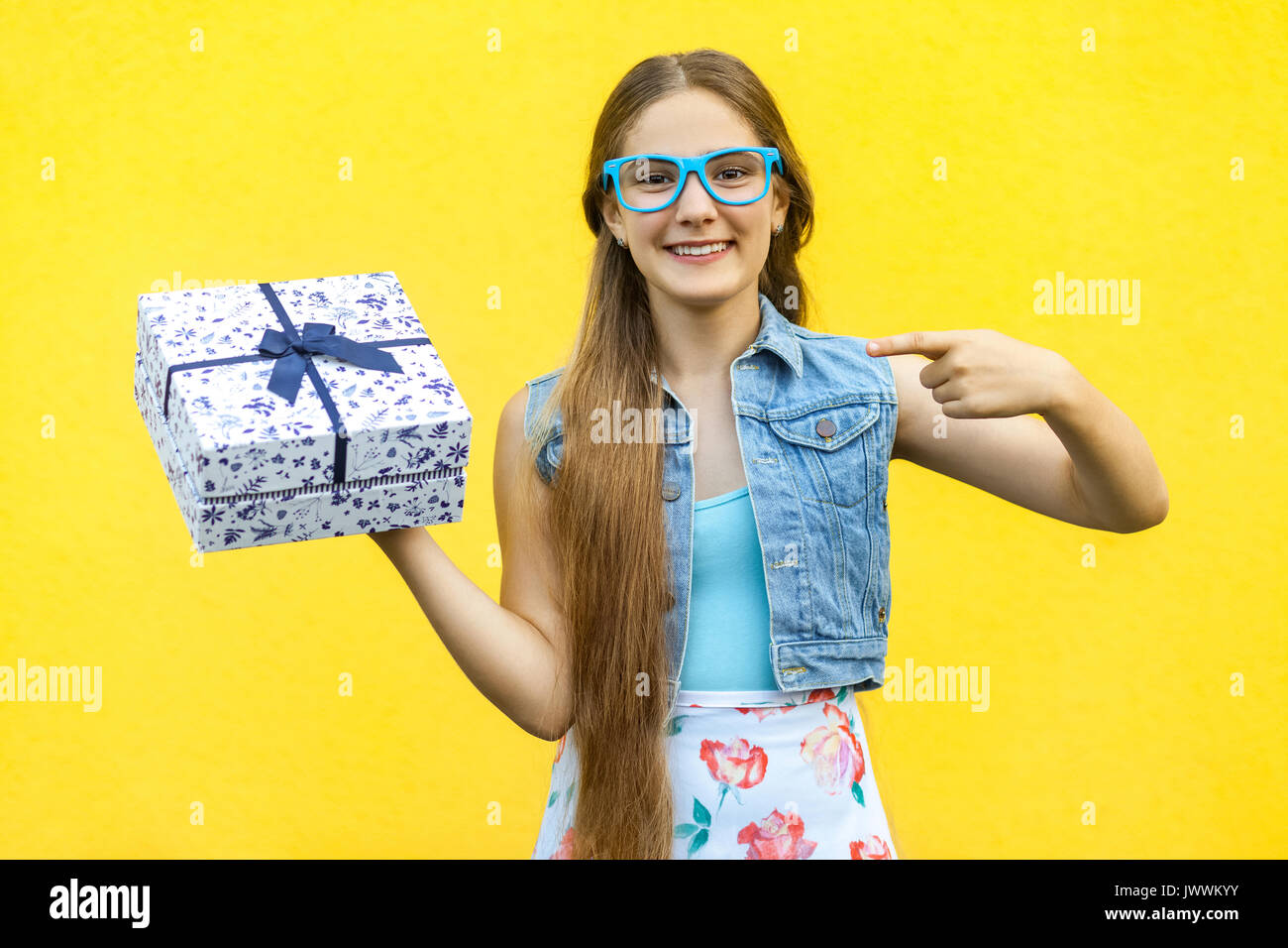 The beautiful smiling girl in dress and blue glasses, pointing finger on present box and toothy smile, isolated over yellow background. Indoor studio  Stock Photo