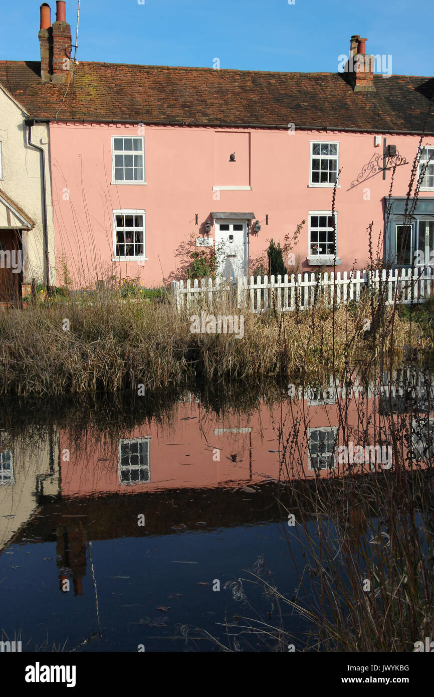 Lovely old pink two story cottage in rural Bushey, Hertfordshire, UK, reflected in the village pond; blue sky, white picket fence and reeds . Stock Photo