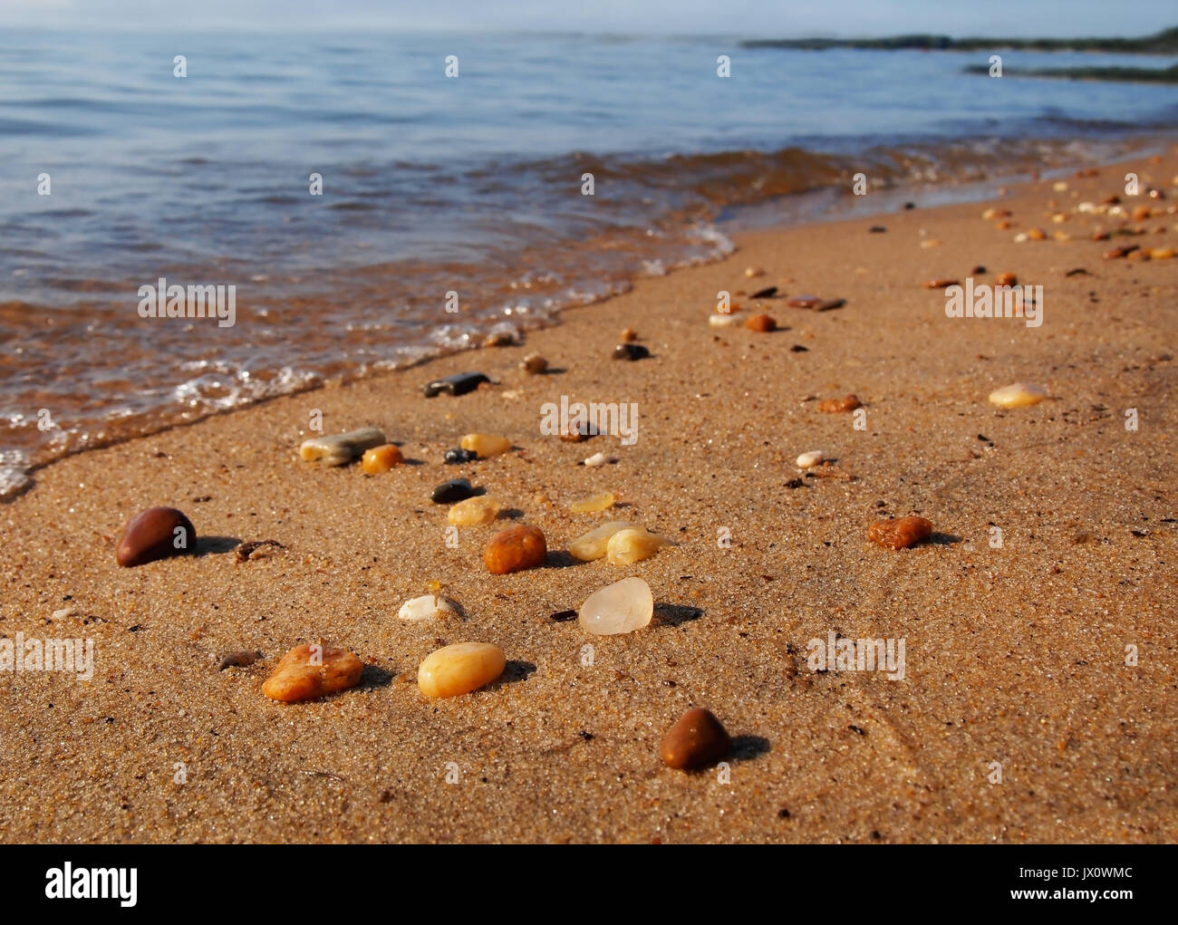 Wet beach pebbles on top of the sand sparkle in the sunlight at the water's edge with gentle, bubbling tides rolling in. Stock Photo