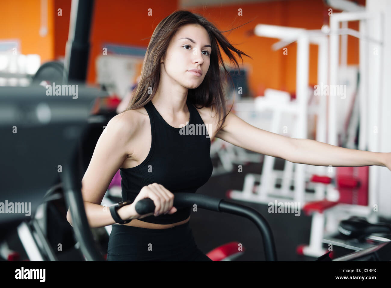 Young beautiful white girl in black sports suit is engaged on a stationary bike in the fitness club. Stock Photo