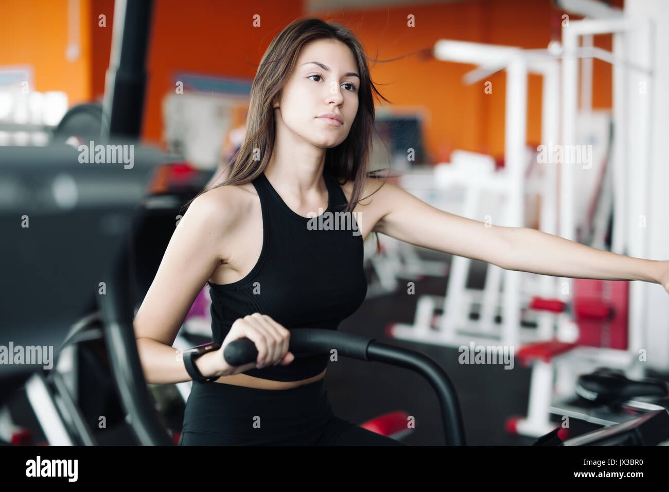 Young beautiful white girl in black sports suit is engaged on a stationary bike in the fitness club. Stock Photo