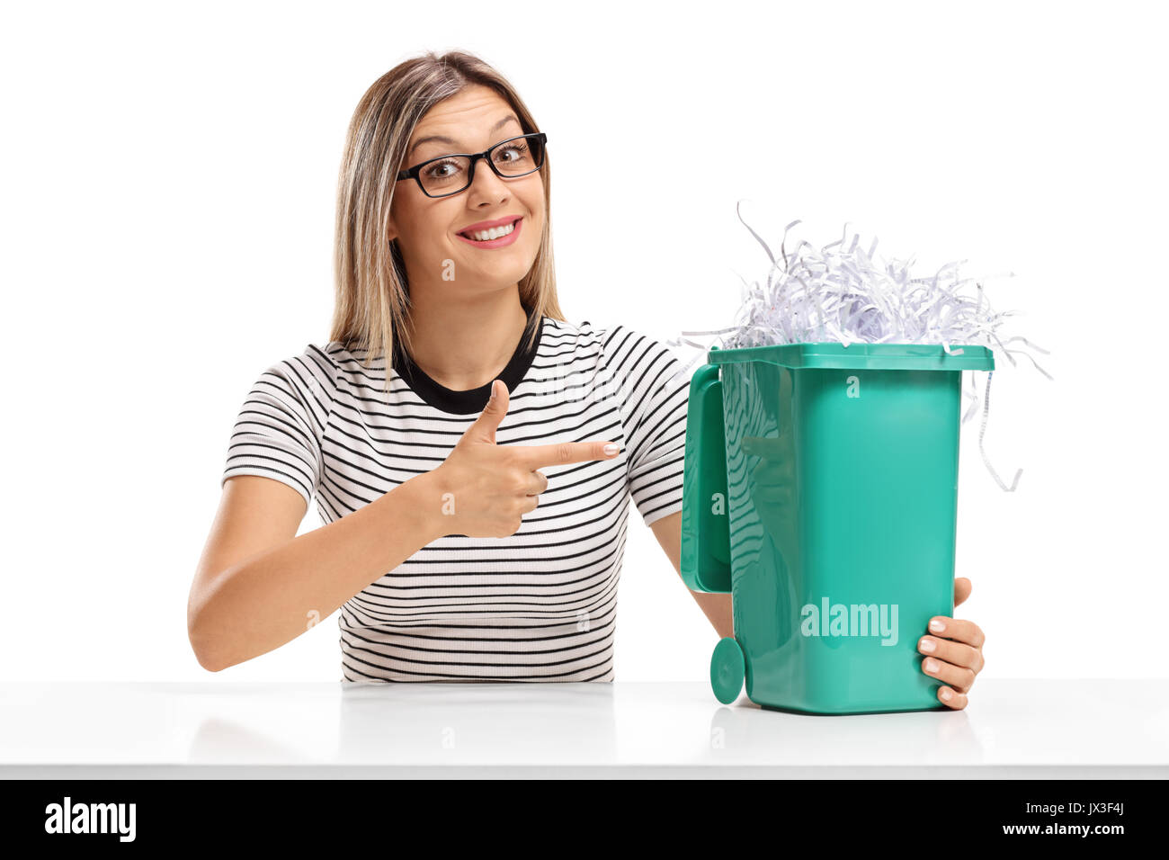 Young woman sitting at a table and pointing at a garbage bin full of shredded paper isolated on white background Stock Photo