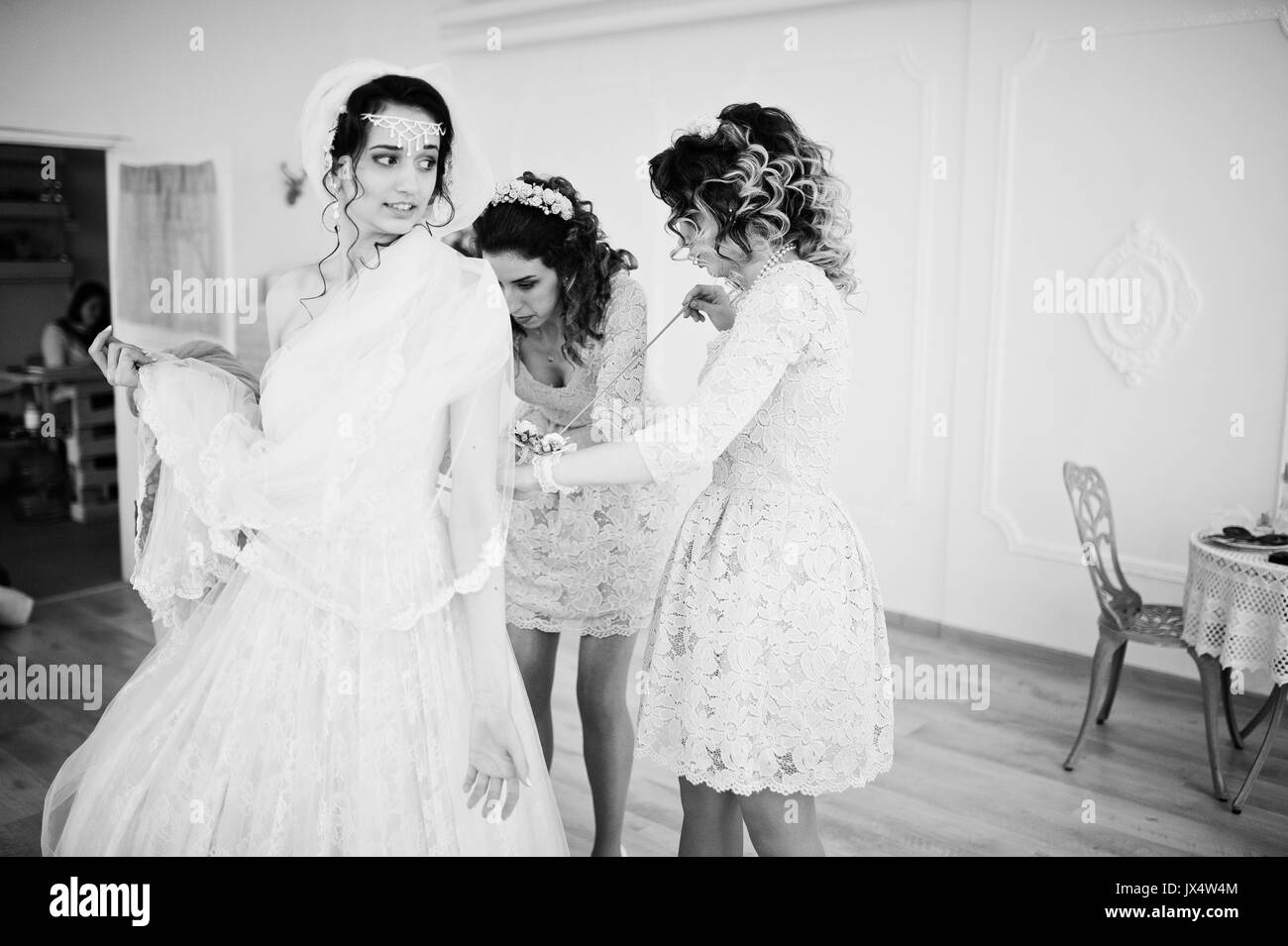 Helpful bridesmaids and mother helping bride to tie the dress up in big light room. Black and white photo. Stock Photo