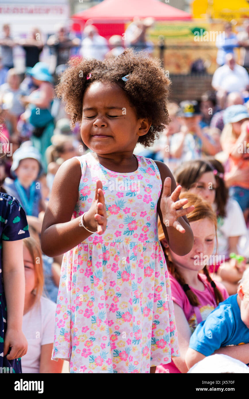Afro-Caribbean child, girl, 6-7 years old, standing up in audience of sitting children, Wears a pink flower dress. Intense facial expression of concentration as she tries to clap. Stock Photo