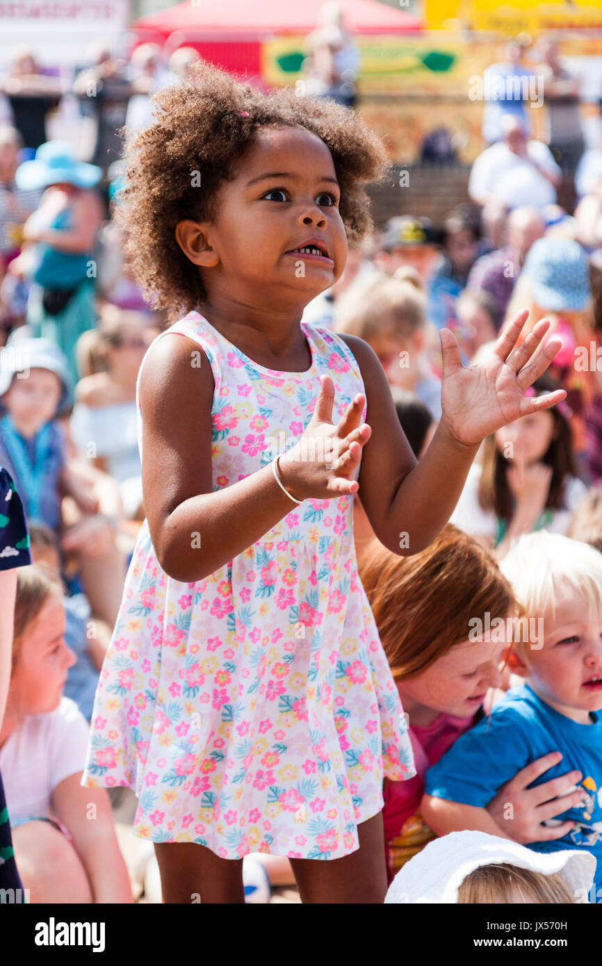 Afro-Caribbean child, girl, 6-7 years old, standing up in audience of sitting children, Wears a pink flower dress. Intense facial expression of concentration as she tries to clap. Stock Photo