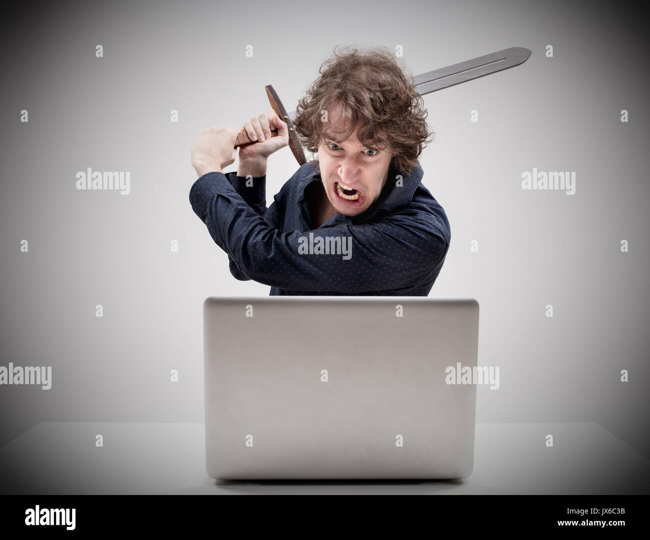 angry man about to destroy his laptop computer by hitting it with a medieval sword with an intense expression of rage - concept of computer hassles an Stock Photo