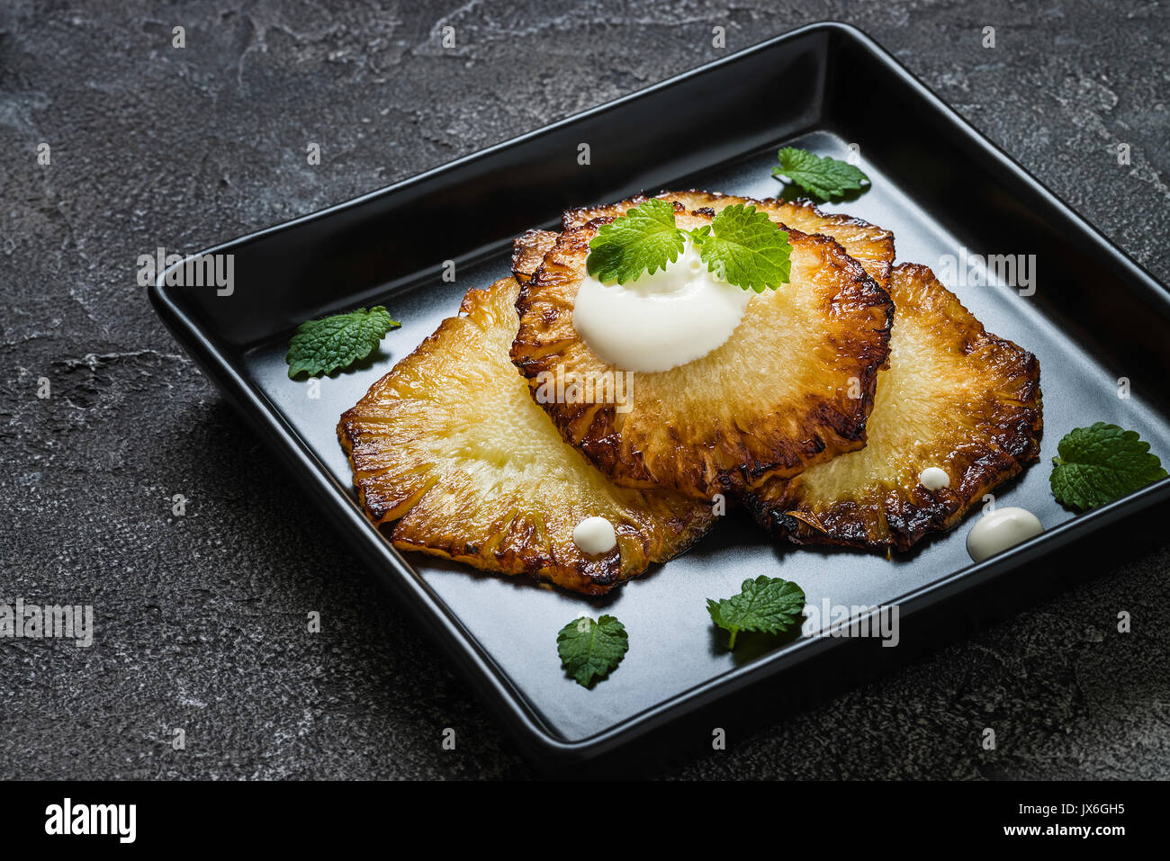 Fried pineapple slices with creme fraiche, mint in black square plate on dark background Stock Photo