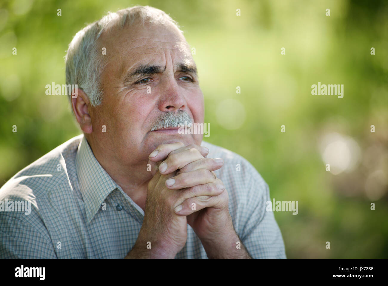 Elderly man sitting thinking in the garden with his chin resting on his hands as he stares into the distance with a pensive expression Stock Photo