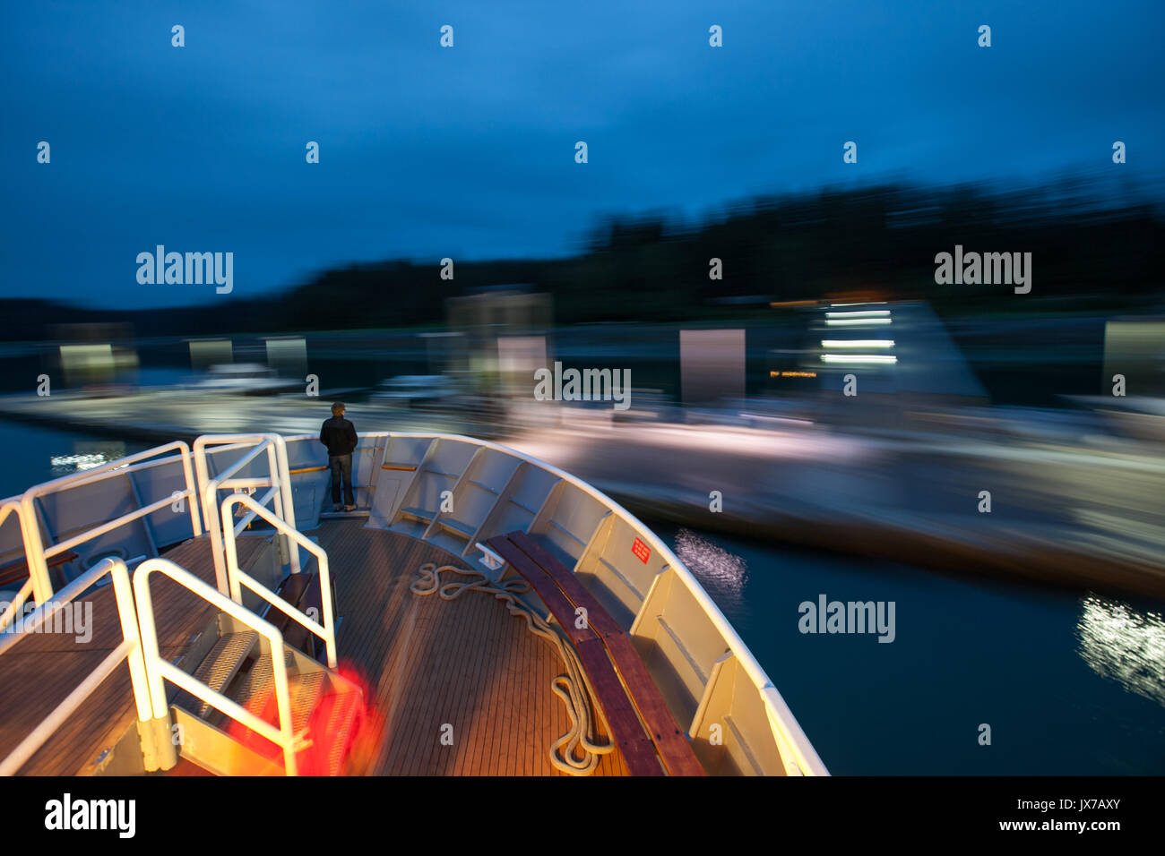 At twilight, a passenger stands on the bow of an expedition cruise ship as it pulls away from the dock. Stock Photo