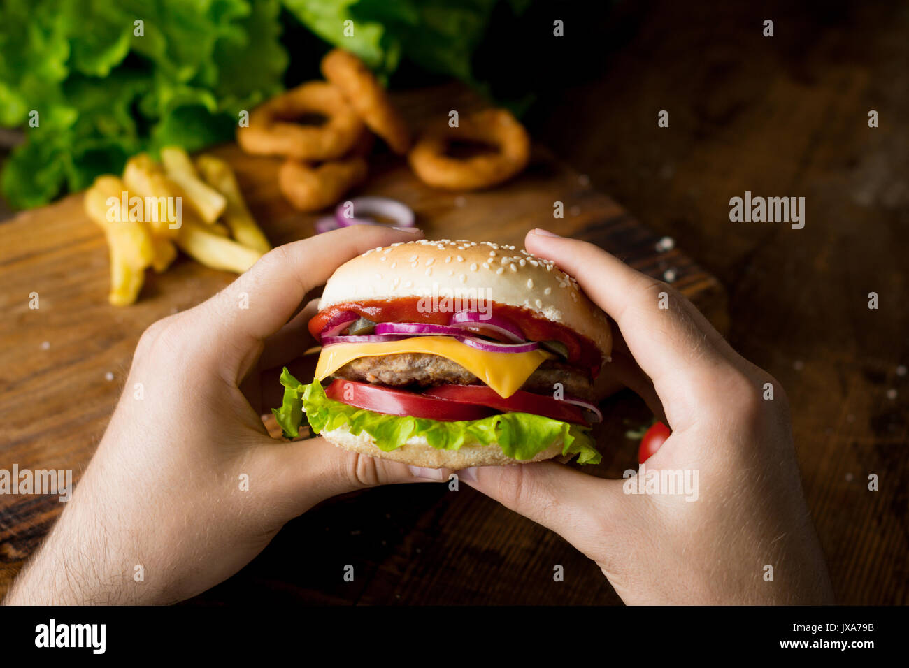 Cheeseburger. Man hands holding burger with cheese, red onion, tomatoes, lettuce green salad and pickles. Square crop. Closeup view, selective focus Stock Photo