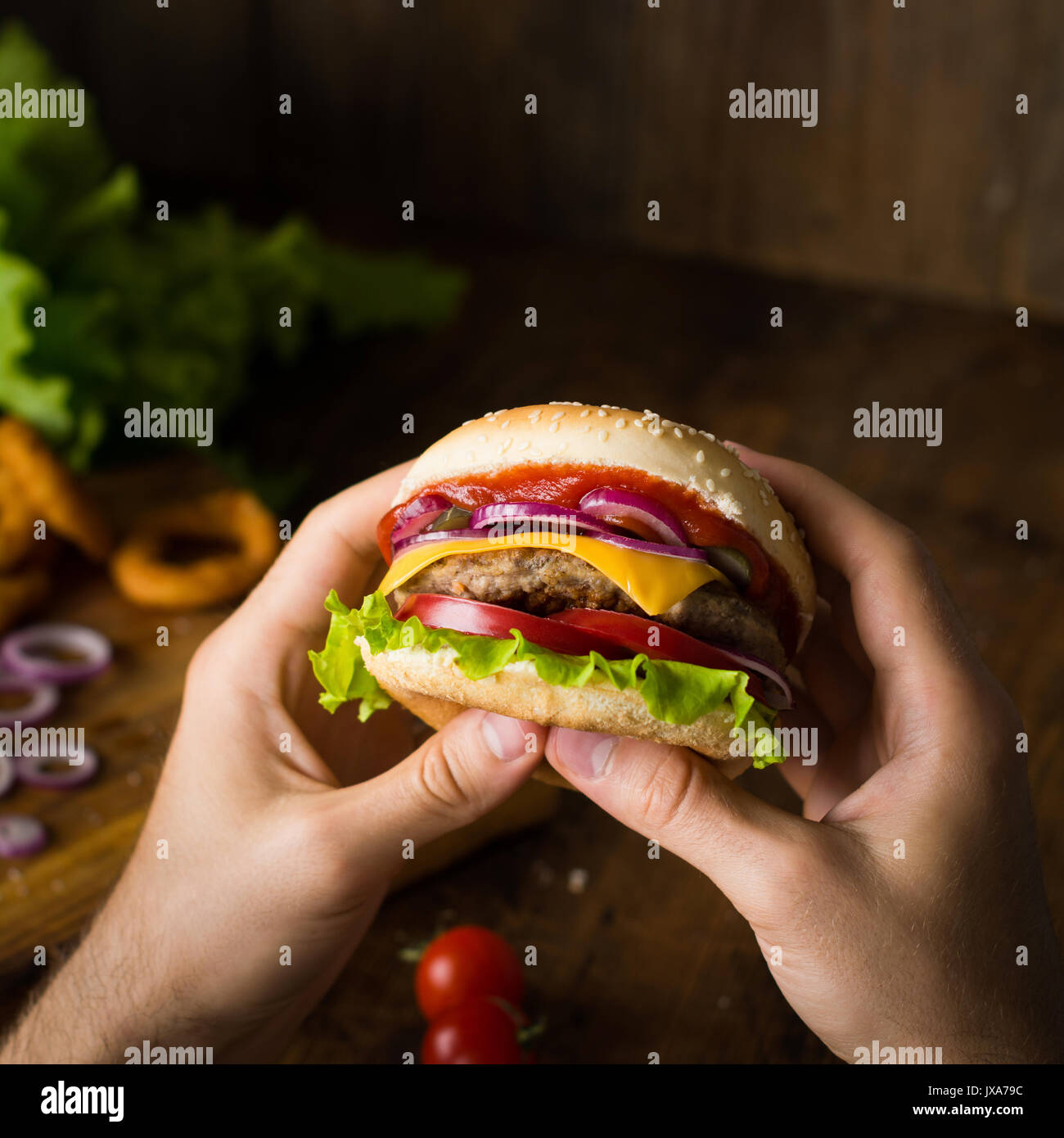 Cheeseburger. Man hands holding burger with cheese, red onion, tomatoes, lettuce green salad and pickles. Square crop. Closeup view, selective focus Stock Photo