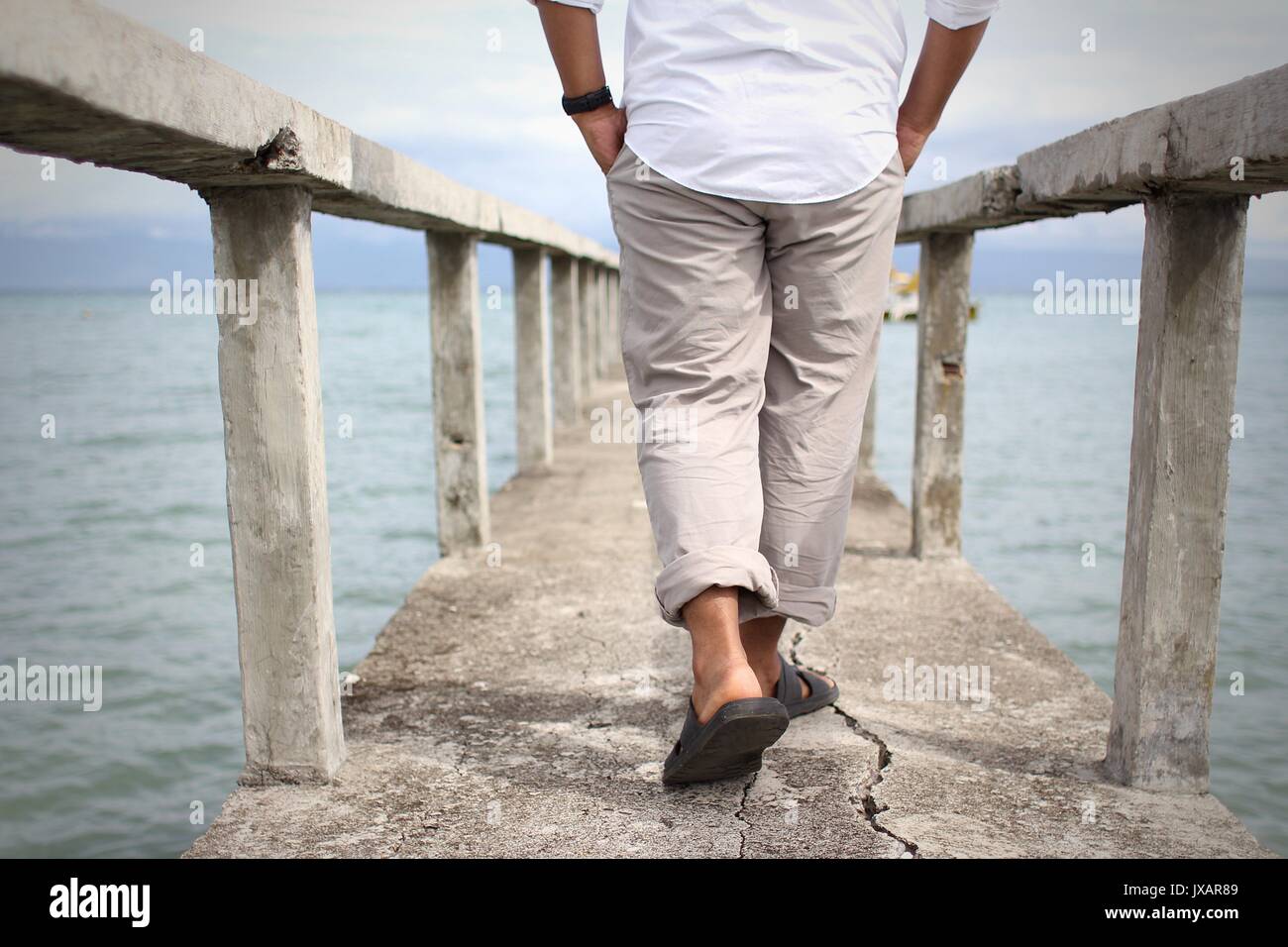 A man walking on a concrete bridge with cracks. The sea and the sky are both visible in the picture. Stock Photo