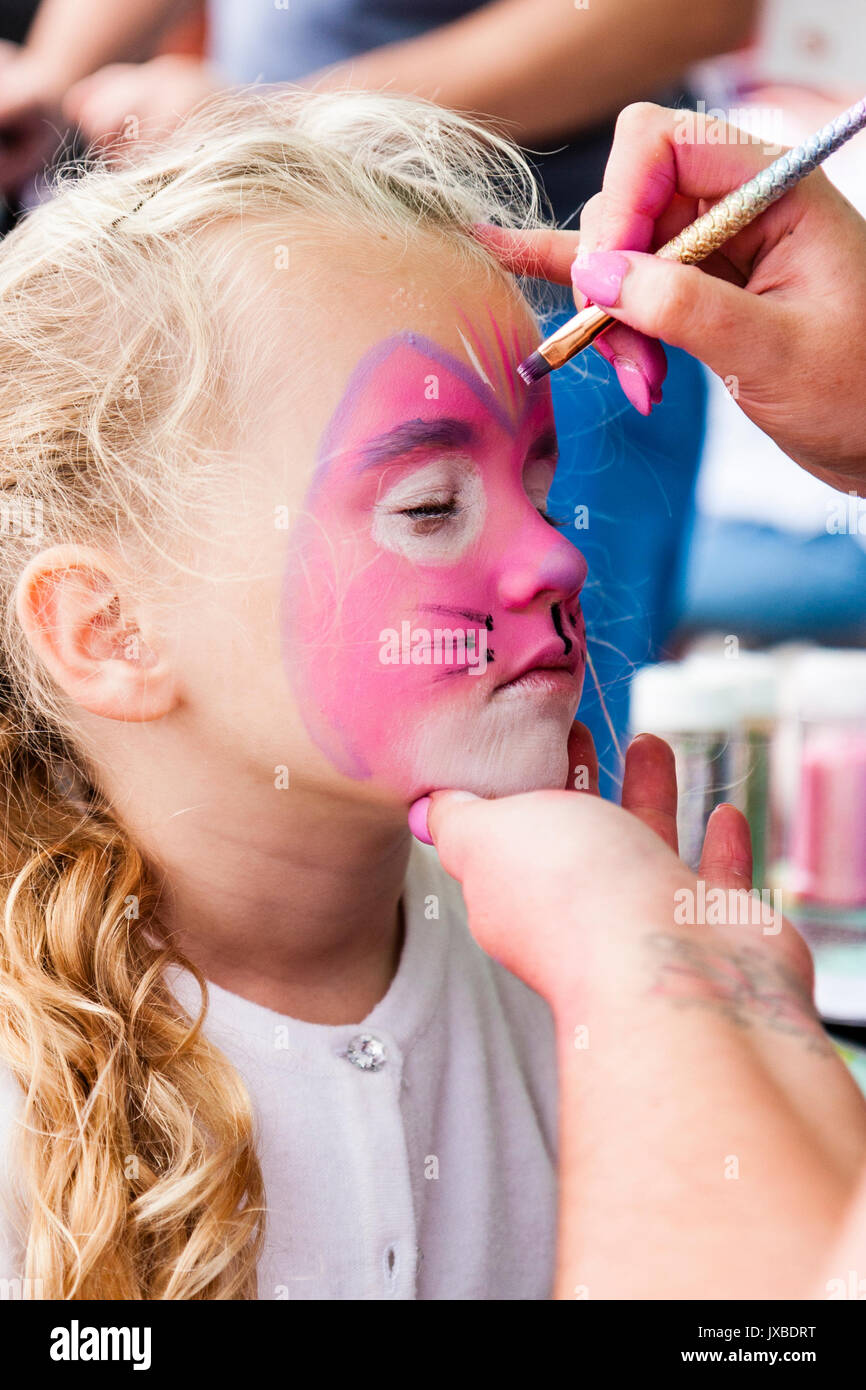 Caucasian blonde child, girl, 7-8 years old, side view, having face painted pink with cat face. Hand holding her chin, another hand holding brush. Stock Photo