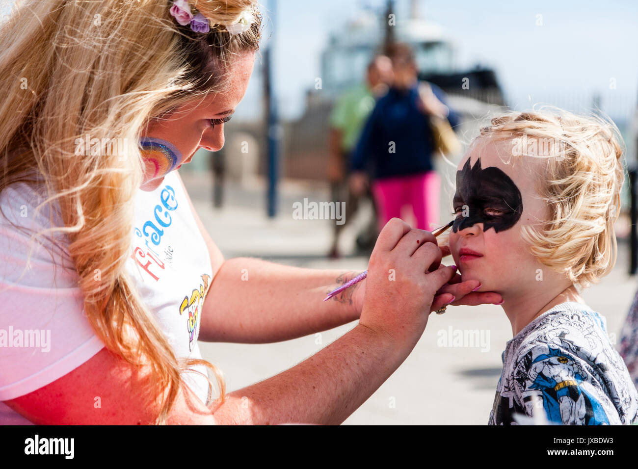 Young Caucasian child, girl, 5-6 years old, sitting in sunshine having her face painted with a batman type mask by blonde woman. Stock Photo