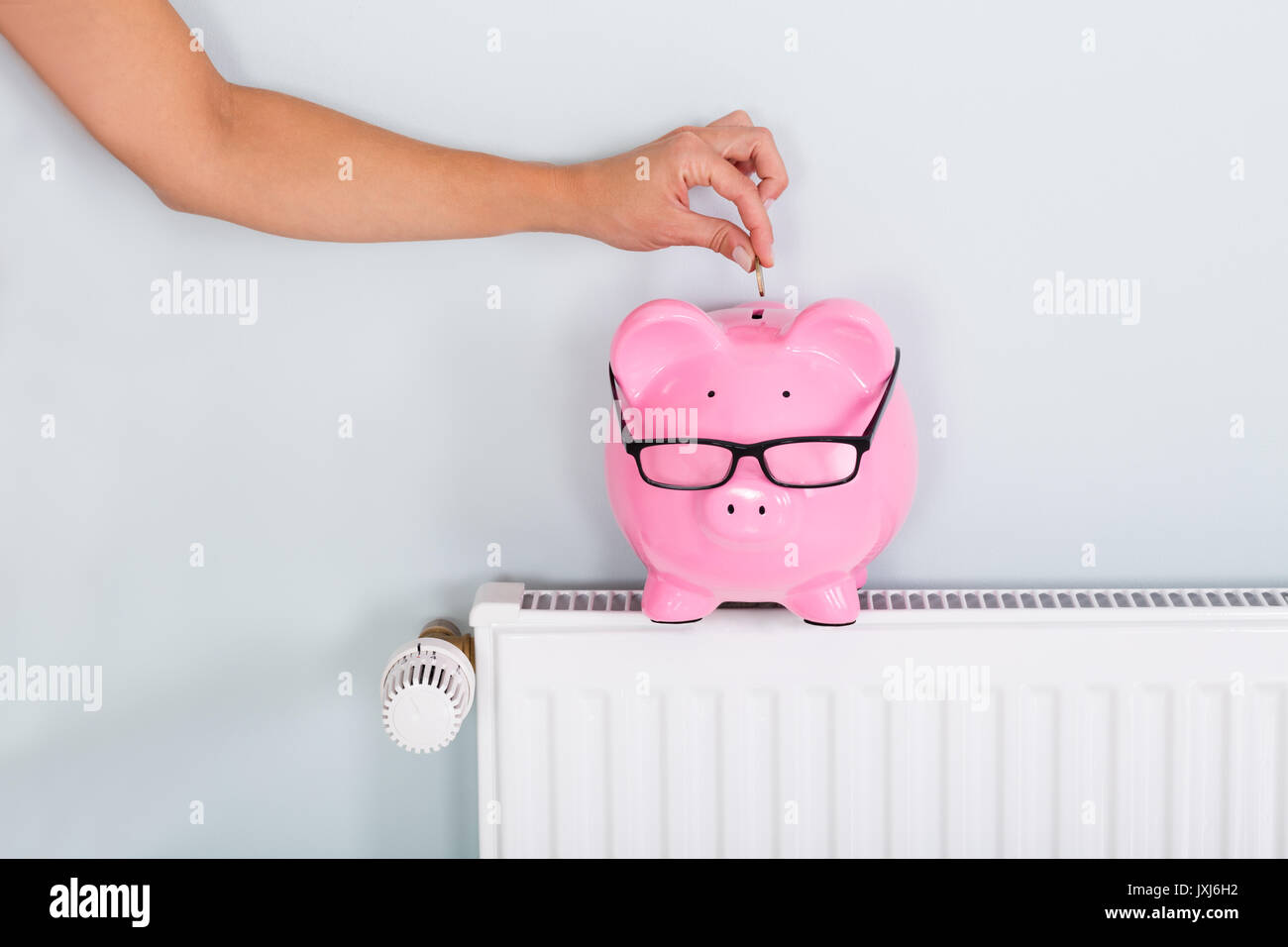 Woman Hand Inserting Coin In Piggy Bank On Radiator To Save On Energy Bill Stock Photo