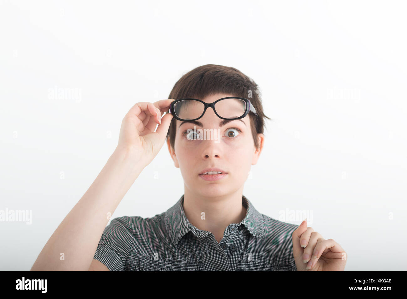 Young beautiful dark-haired woman in grey shirt with astonishment looks upwards, against white studio background. Breaking news. Surprised pleasant woman holding his glasses on forehead Stock Photo