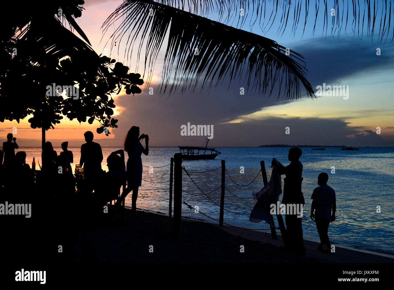 TANZANIA Zanzibar, Stone town, Livingstone beach restaurant and bar, evening at sea, foreign tourist in bar, woman taking a photo of sunset and street hawker selling clothes Stock Photo