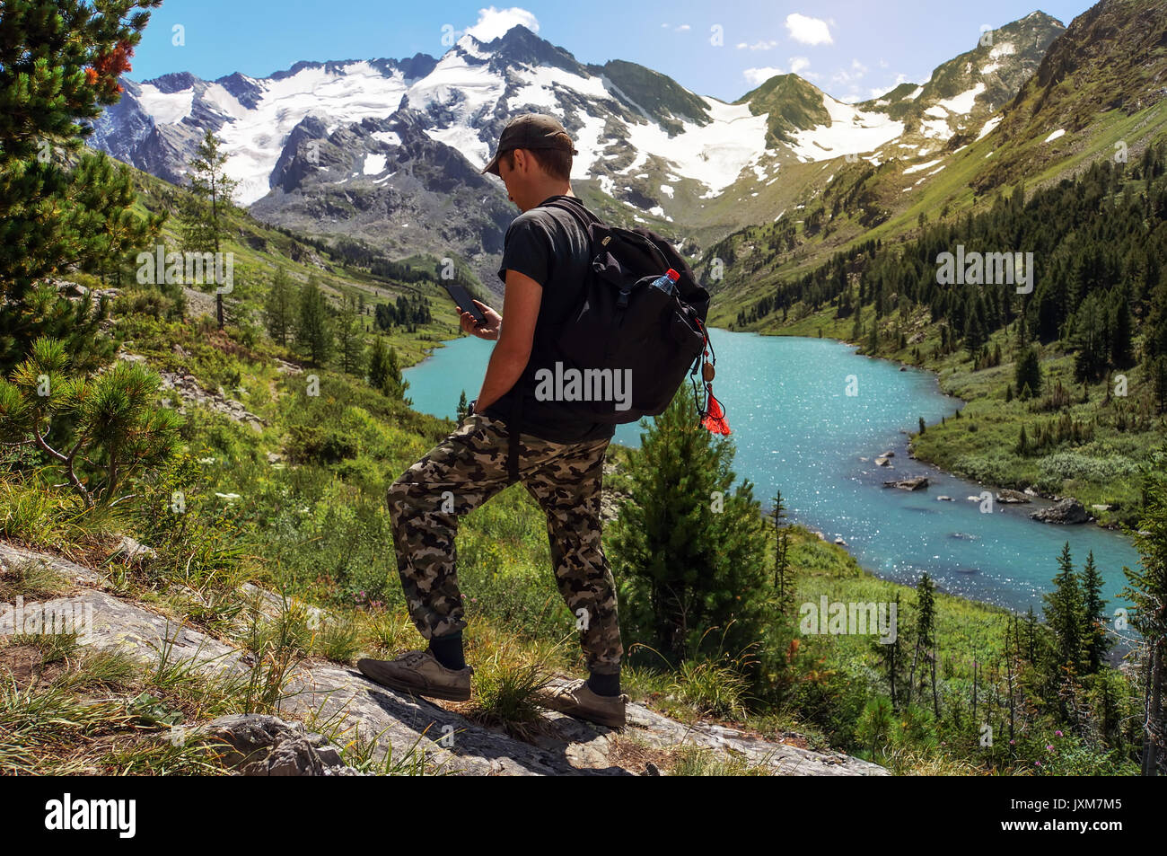 Hiker with backpack standing on a hillside and looking in the navigation on smartphone in the background beautiful mountain landscape with turquoise l Stock Photo