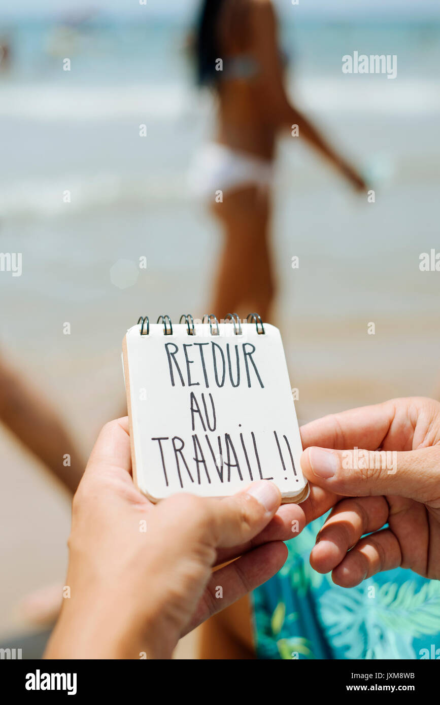 closeup of a young man sitting in a deck chair in the seashore of a beach showing a spiral notepad with the text retour au travail, back to work in Fr Stock Photo
