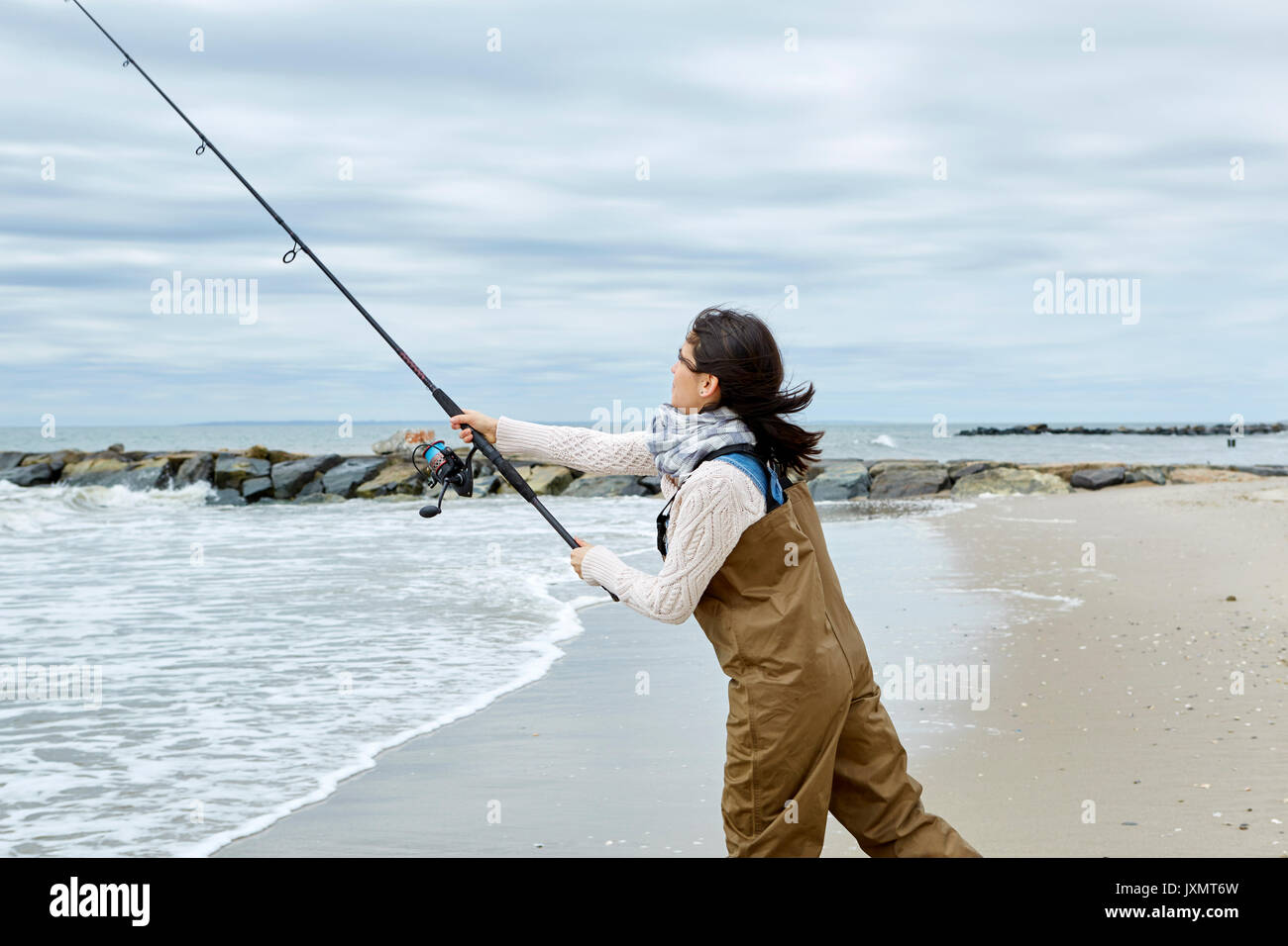 Young woman in waders casting sea fishing rod from beach Stock Photo