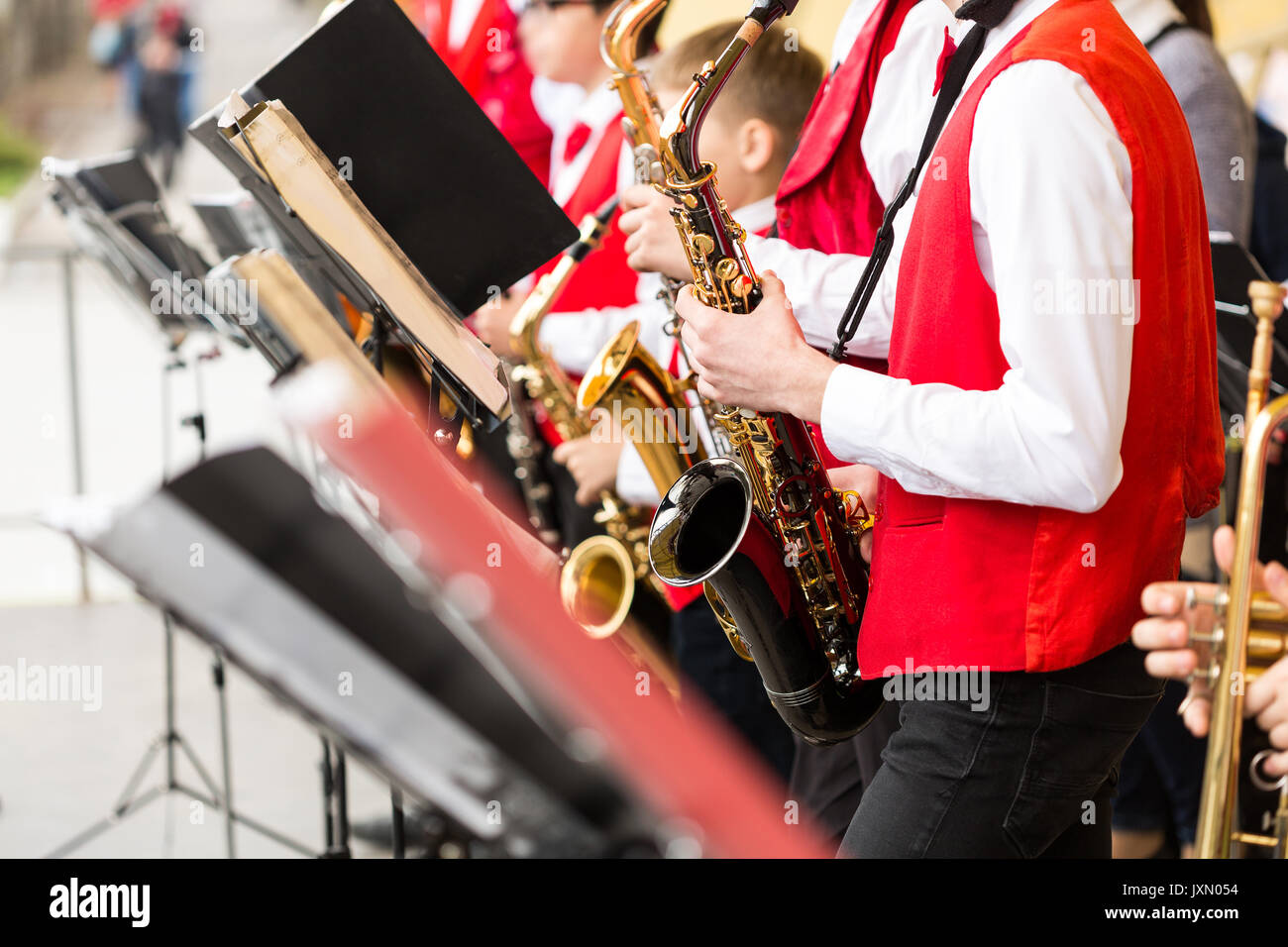 musical instrument, brass band and orchestra concept - festive performance of saxophones, closeup on sax and sheet music stands, musicians in red concert jackets and shirts, selective focus. Stock Photo