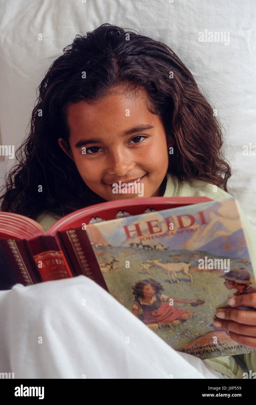 Little girl 7-9 years old with sincere expression reading classic storybook 'Heidi'  in bed looks up open welcoming smile. African American/Caucasian Stock Photo