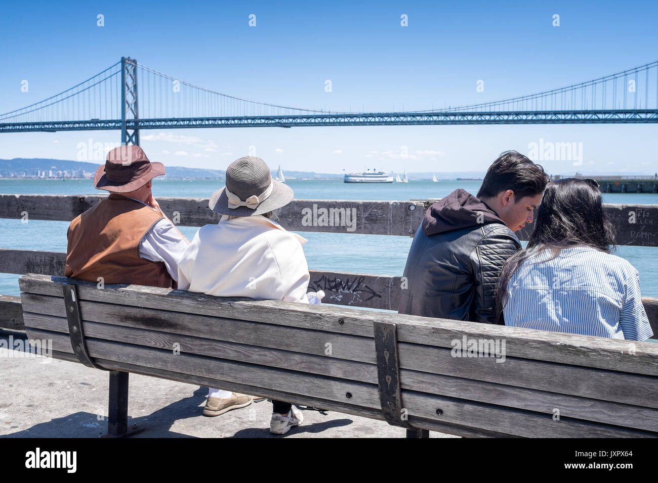 Couples sit on a bench with a view of the Oakland Bay Bridge in the background. One young couple, one senior. Location: Ferry Building, San Francisco Stock Photo
