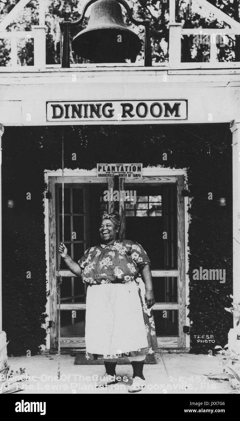 Full-body portrait of African American woman, standing in front of a building titled 'Dining Room, ' wearing a patterned dress and an apron, clutching a rope, with a smiling facial expression, 1920. Stock Photo