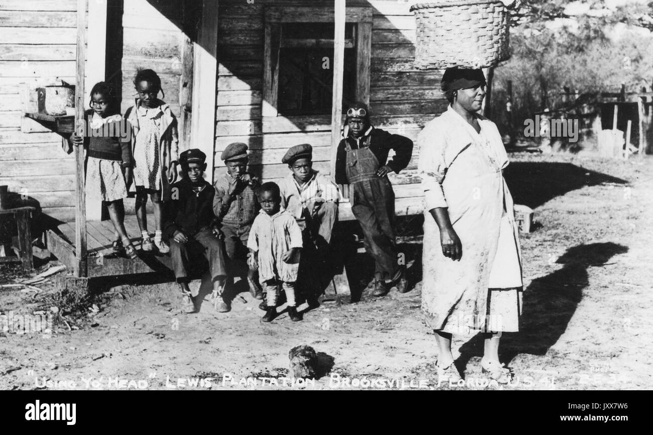 A group of seven young African American children with serious expressions are gathered on the porch of a shanty on the Lewis Plantation in Brooksville, Florida, as a mature African American woman with a neutral expression carries a basket on her head, Florida, 1940. Stock Photo