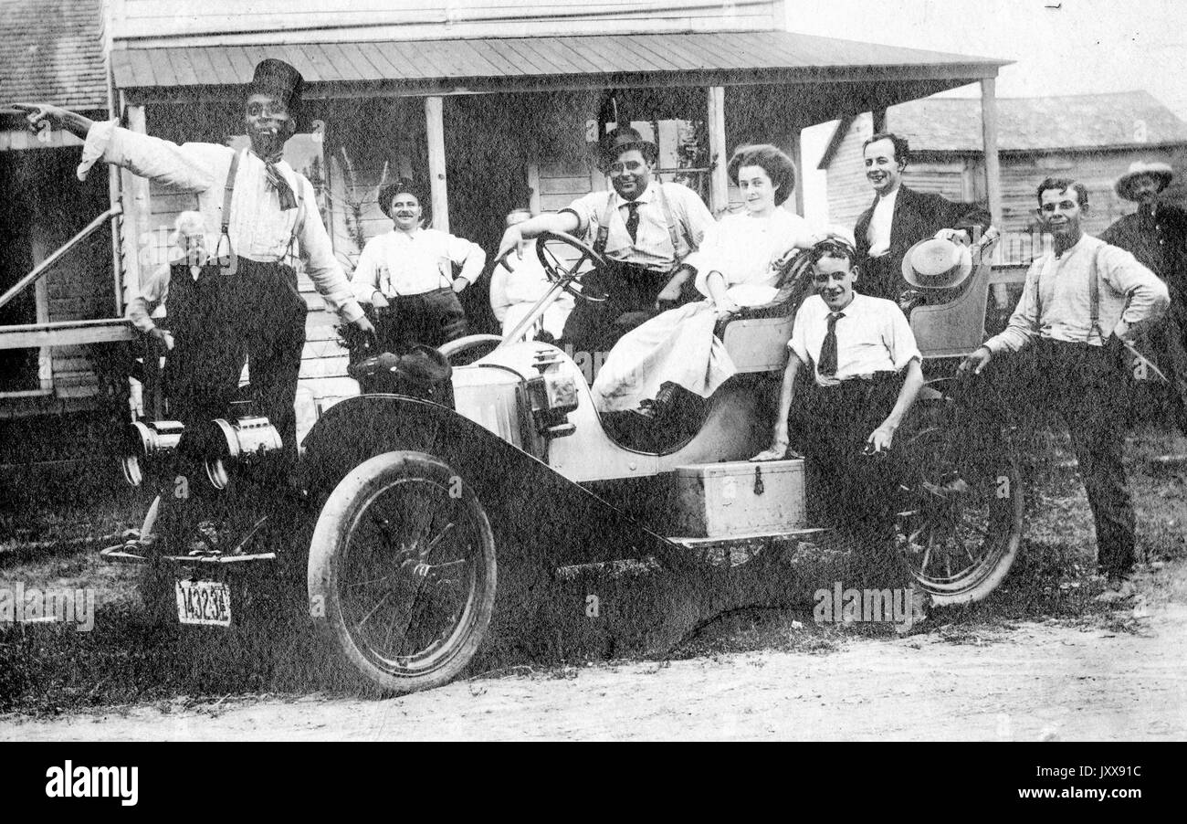 A group of people, including eight men and two women, mostly smiling, stand on or around a car, with one African American man wearing a tophat standing on the fender and pointing with his arm outstretched, 1910. Stock Photo