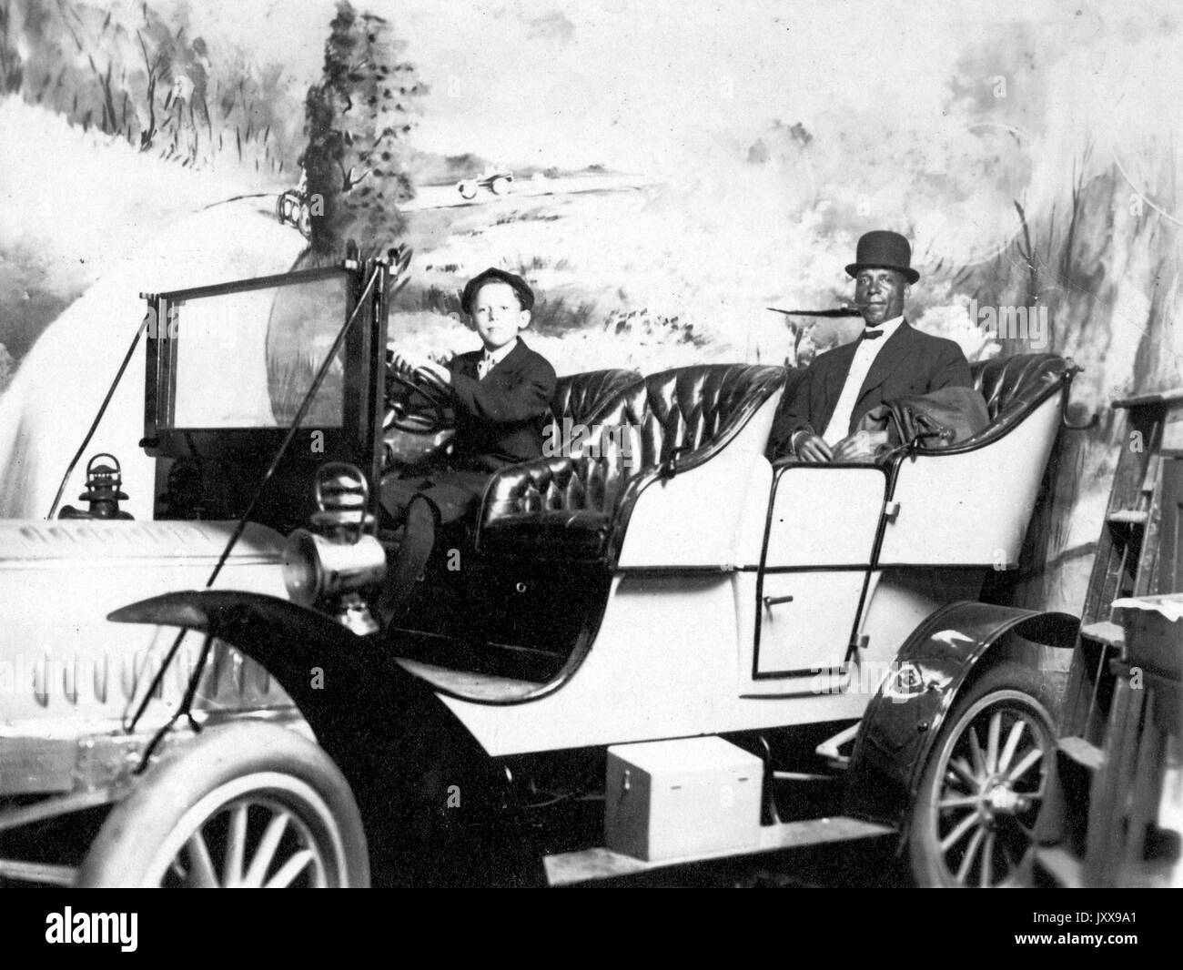 Full length portrait of two men in a car, a young Caucasian boy at the wheel, an African American man in the backseat, wearing a suit and hat, smiling facial expressions, 1915. Stock Photo