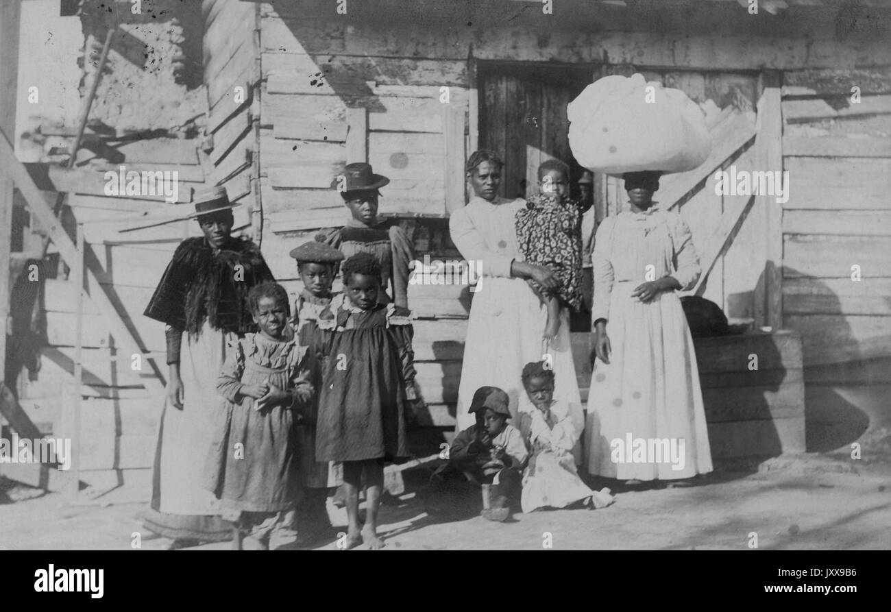 Full length portrait of an African American family: one mature woman wearing a hat and dress, three young girls wearing dresses, one teenage girl in a hat and dress, two children seated by a woman's feet holding a baby, another woman in a white dress and large white hat, neutral facial expressions; Boston, Massachusetts, 1915. Stock Photo