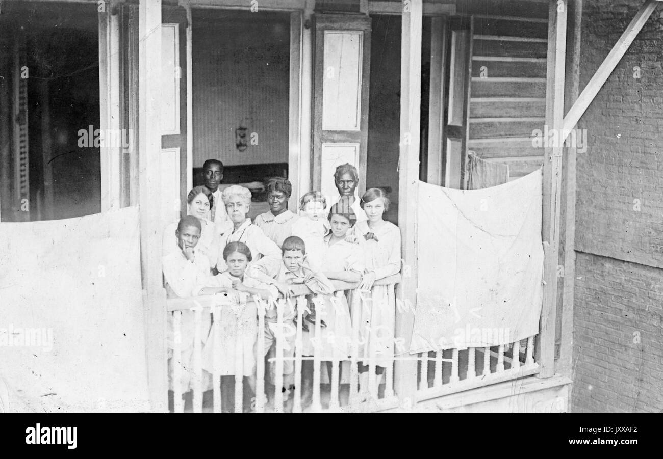 Two mature African American women, one young African American male, one young African American male child, one young African American female child, one elderly white woman, one young white woman, three young white female children, one young white male child all standing on porch of house, all wearing light clothing, 1907. Stock Photo